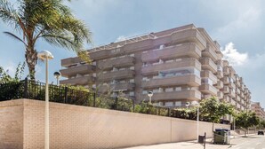 Un edificio residencial de varios pisos con balcones, un muro de ladrillo y una calle con un contenedor de basura y un farol.