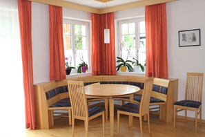 Un coin repas comprenant une table ronde et six chaises, un banc intégré et de grandes fenêtres avec des rideaux.