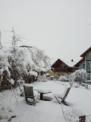 Zona exterior cubierta de nieve con un banco y una mesa, rodeada de arbustos y árboles cargados de nieve.
