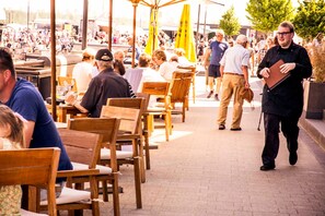 Un espacio exterior para comer con mesas y sillas de madera, sombrillas amarillas y mucha gente.