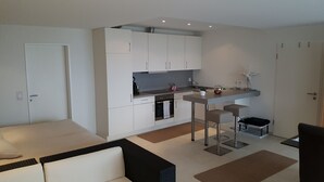 Una cocina moderna con armarios blancos, horno empotrado y un pequeño comedor con dos taburetes.