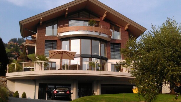 Una casa moderna de dos pisos con un balcón curvo, ventanas amplias y un automóvil estacionado en la entrada.