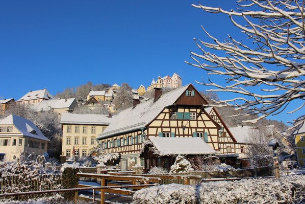 A traditional half-timbered building with a snow-covered roof, surrounded by other buildings and a clear blue sky. - Category A: 1-room apartment for 2 persons - Pension Mühle (Egloffstein)