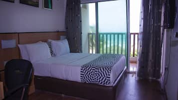 A bedroom with a bed, a chair, a wooden headboard, a balcony with a view, and a TV mounted on the wall.