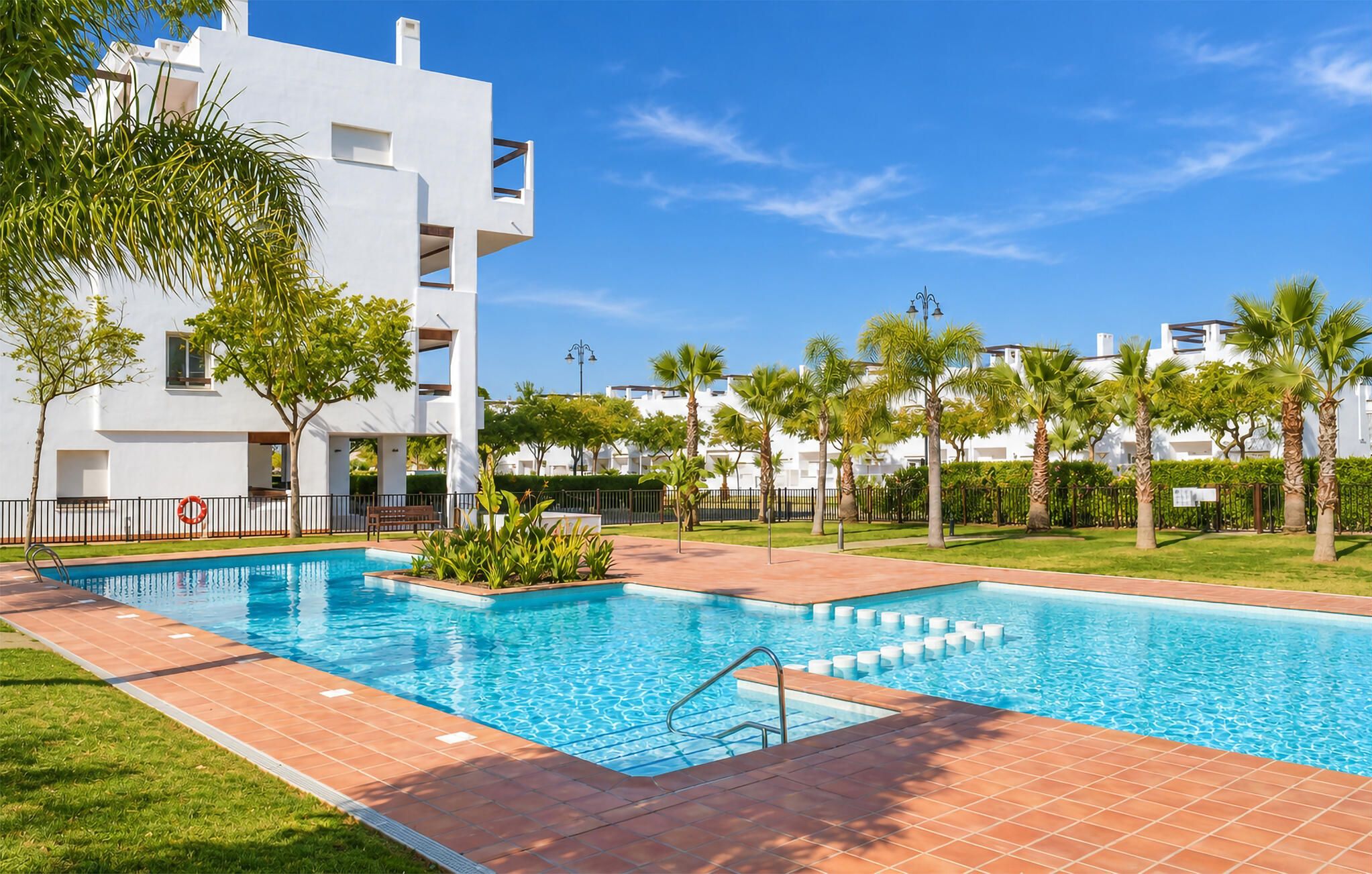 Área de piscina com céu claro, rodeada por palmeiras e um edifício branco.