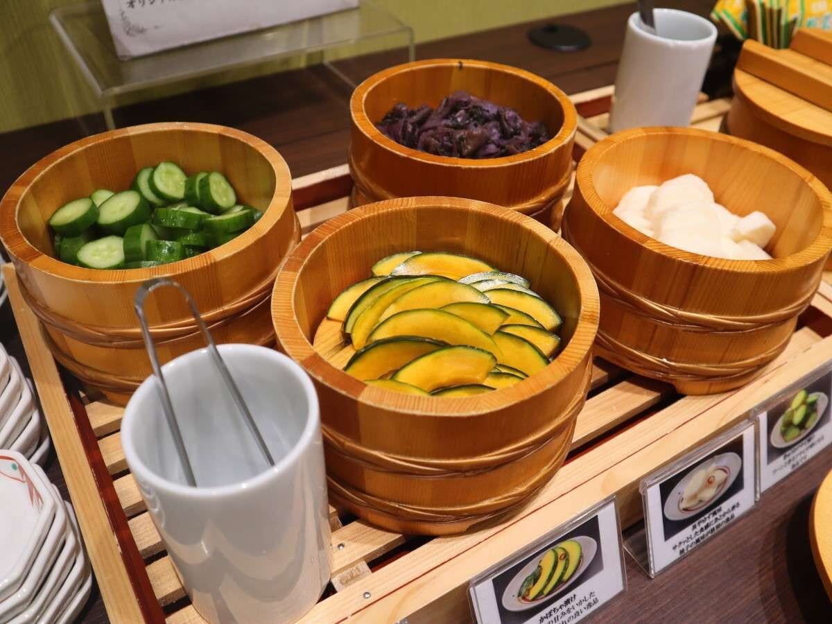 Un bufé con cuencos de madera que contienen pepinos en rodajas, calabacín amarillo, verduras moradas encurtidas y queso blanco.