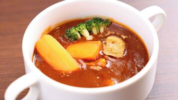 Una taza de cerámica blanca con asas, que contiene una sopa con trozos visibles de zanahoria y brócoli, y un trozo de tofu.