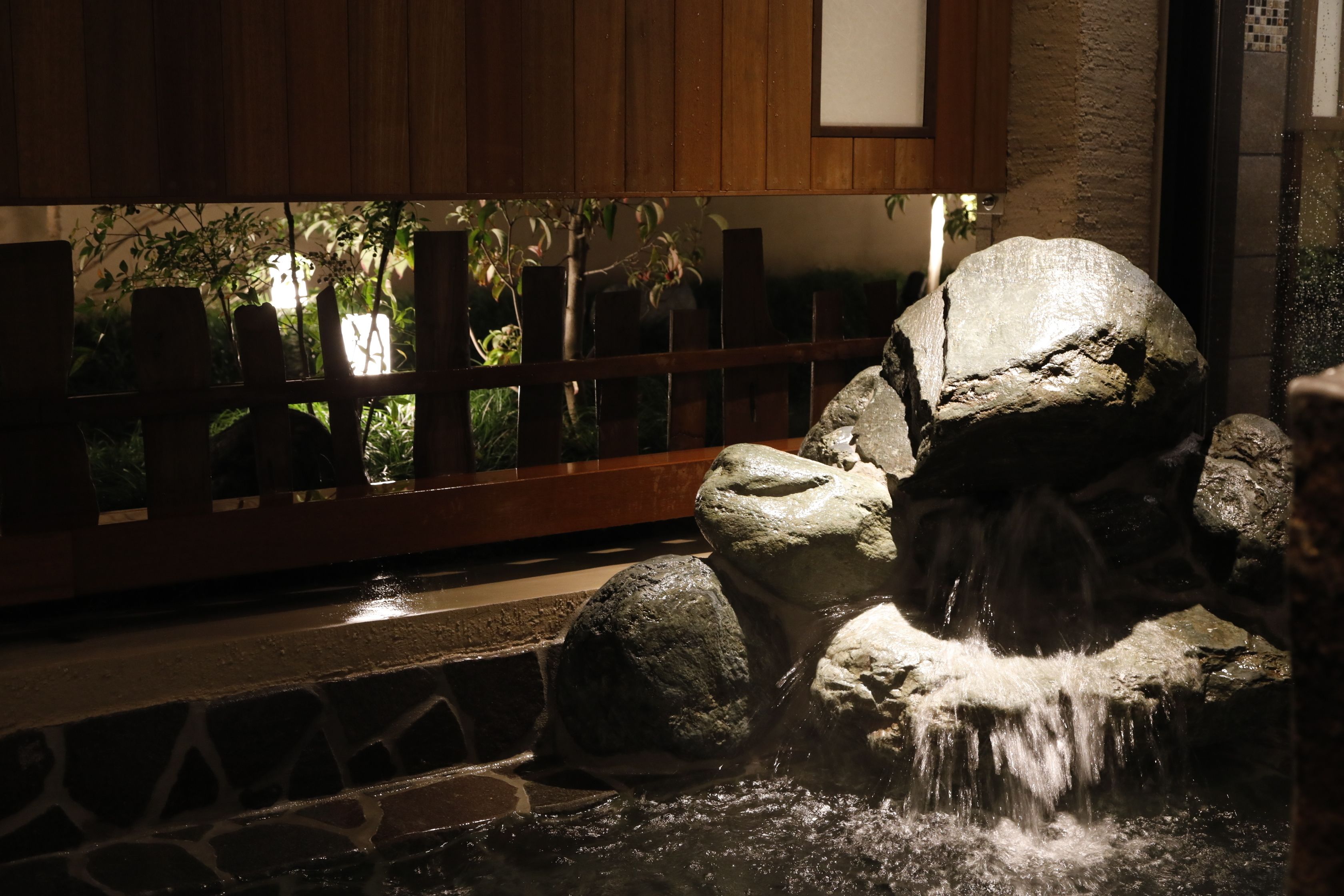 A stone water feature with a waterfall, surrounded by a wooden fence and a stone wall.