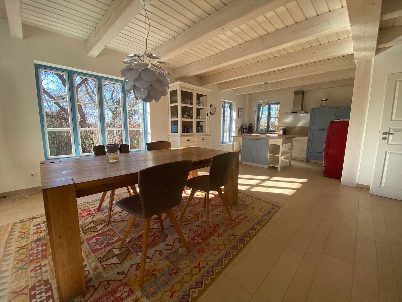 A spacious kitchen with a wooden table and chairs, a red refrigerator, and a view of the outside through large windows.