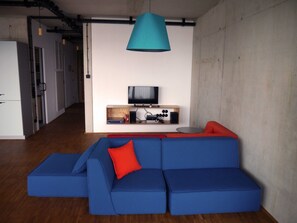 A modern living room with a blue sectional sofa, a red accent pillow, a teal pendant light, a flat-screen TV, and a wooden shelf with books and electronic devices. - The Playful (11) - Z1-THE HOUSE (Zwenkau)