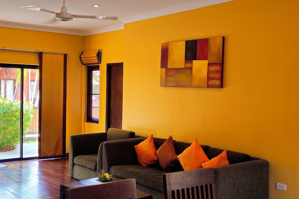 A room with a yellow wall, a grey sofa with orange cushions, a wooden chair, and a ceiling fan.
