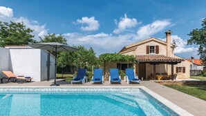A swimming pool with lounge chairs, a covered patio, and a traditional house with a tiled roof.