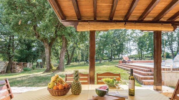 An outdoor dining area with a table set for two, featuring a fruit basket, a pineapple, a watermelon, and a bottle of wine.