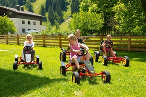 Four children riding toy tractors on a grassy field with a wooden fence and a building in the background. - Rauriser Jungle Family Studio - Hotel Alpina **** (Rauris)