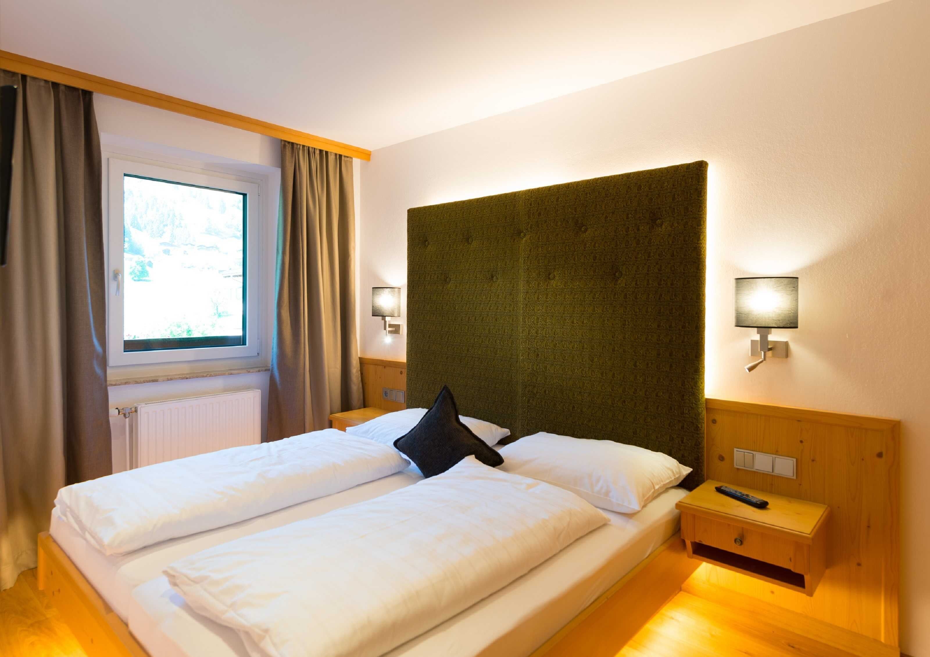 A double bed with white linens, a dark headboard, two bedside lamps, a window with curtains, and a wooden nightstand with a remote control.