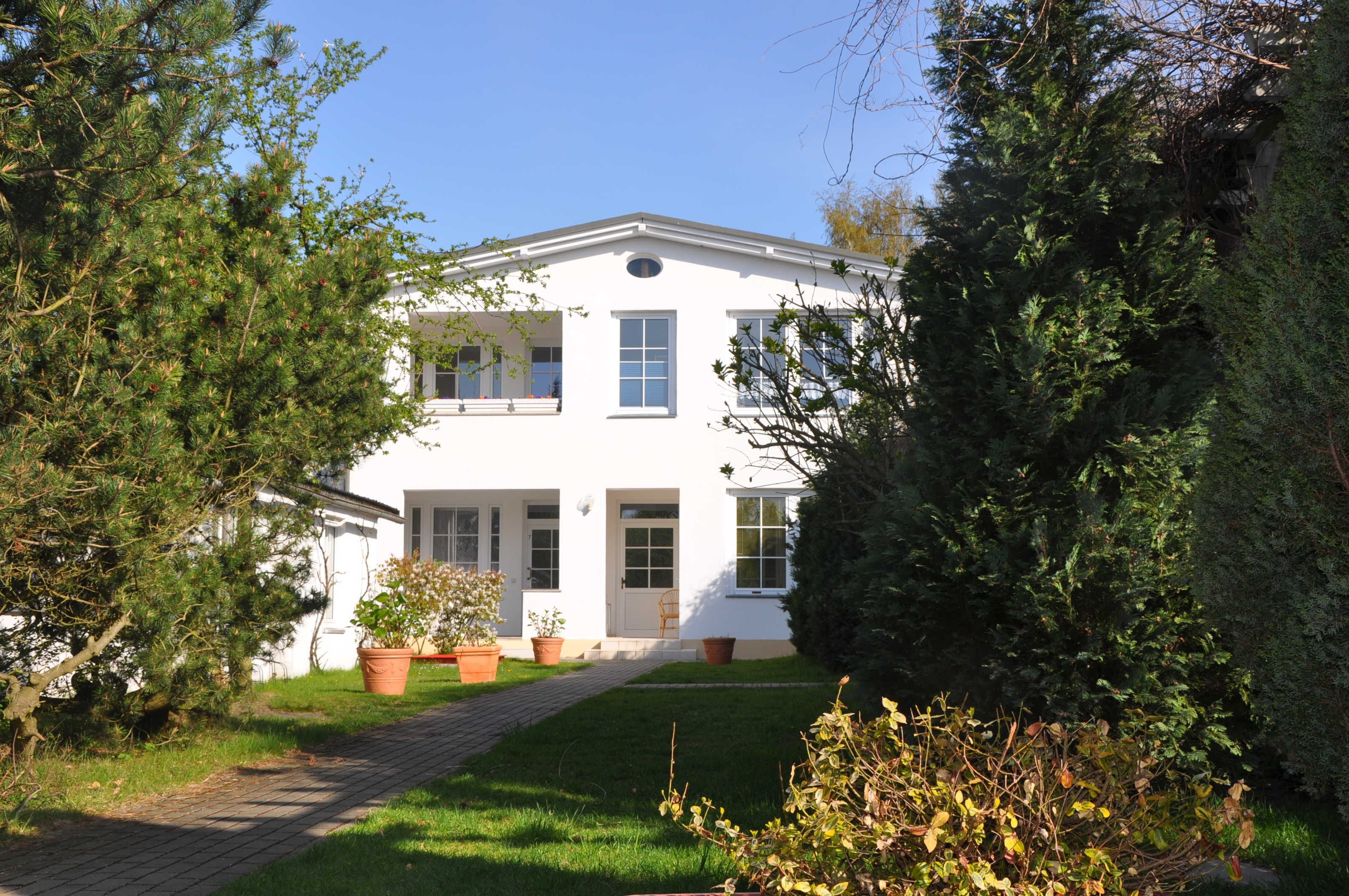 A white two-story building with a paved pathway leading to the entrance, surrounded by greenery.