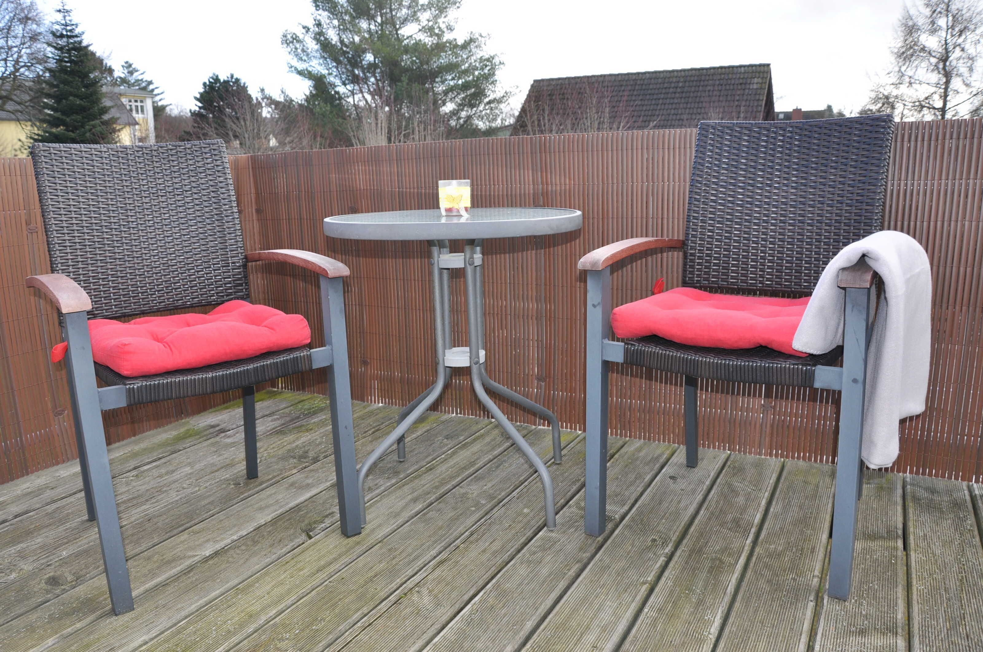 Two wicker chairs with red cushions and a small round table with a candle on it, set on a wooden deck.