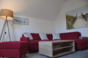 Um sofá vermelho com almofadas estampadas, uma mesa de centro de madeira, um abajur e uma pintura com tema de praia na parede.