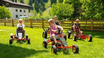 Des enfants montent de petits tracteurs jouets dans un champ d’herbe, avec une clôture en bois et un bâtiment en arrière-plan.