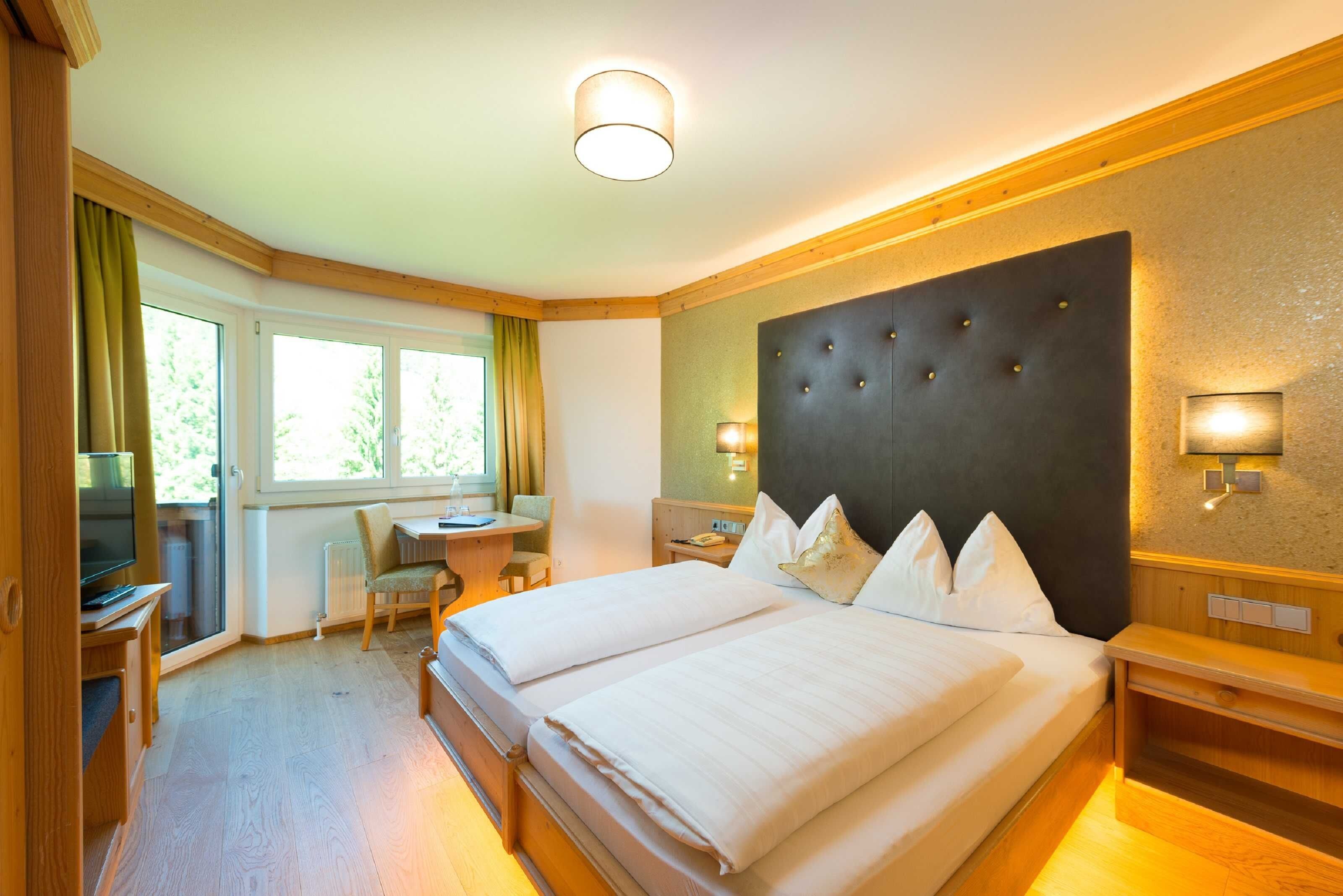 Una habitación de hotel con una cama grande, un escritorio, una silla, un televisor y un ventanal con cortinas.
