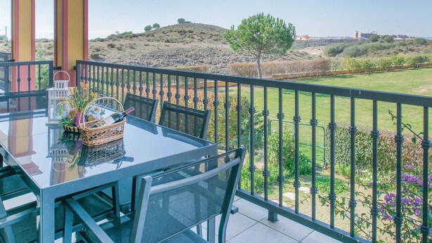 Ein Balkon mit Tisch und Stühlen, der auf eine hügelige Landschaft mit spärlicher Vegetation und weit entfernte Gebäude blickt.