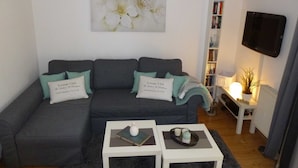 Een woonkamer met een grijze bank, twee witte bijzettafeltjes, een lijstwerk met bloemen, een boekenkast en een aan de muur bevestigde televisie.