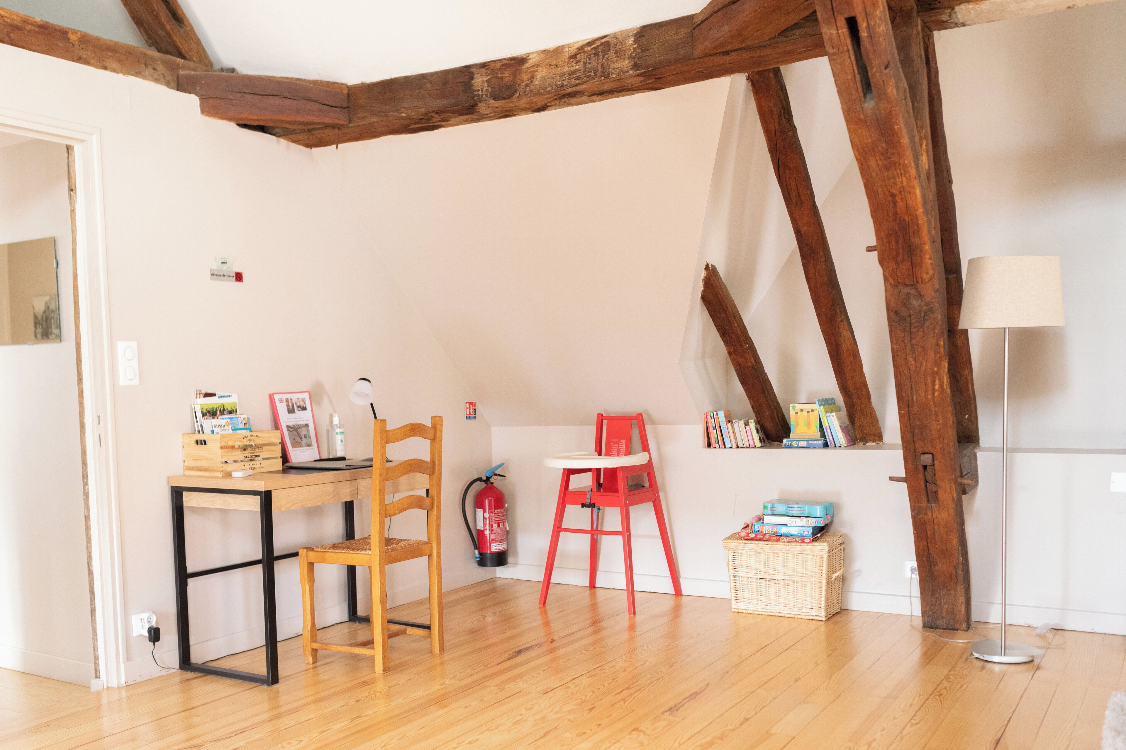 Une pièce avec des poutres en bois, un bureau avec une chaise, une chaise haute rouge, un lampadaire et un panier rempli de livres.