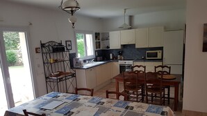 Une cuisine avec des meubles blancs, une table à manger recouverte d’une nappe à motifs et un luminaire suspendu.