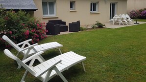 Une maison avec un toit en tuiles, une pelouse et du mobilier d’extérieur comprenant des chaises longues blanches et une table.