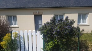 Une maison avec une clôture en bois peinte en blanc, un petit jardin avec différentes plantes et une clôture en métal à l’avant.