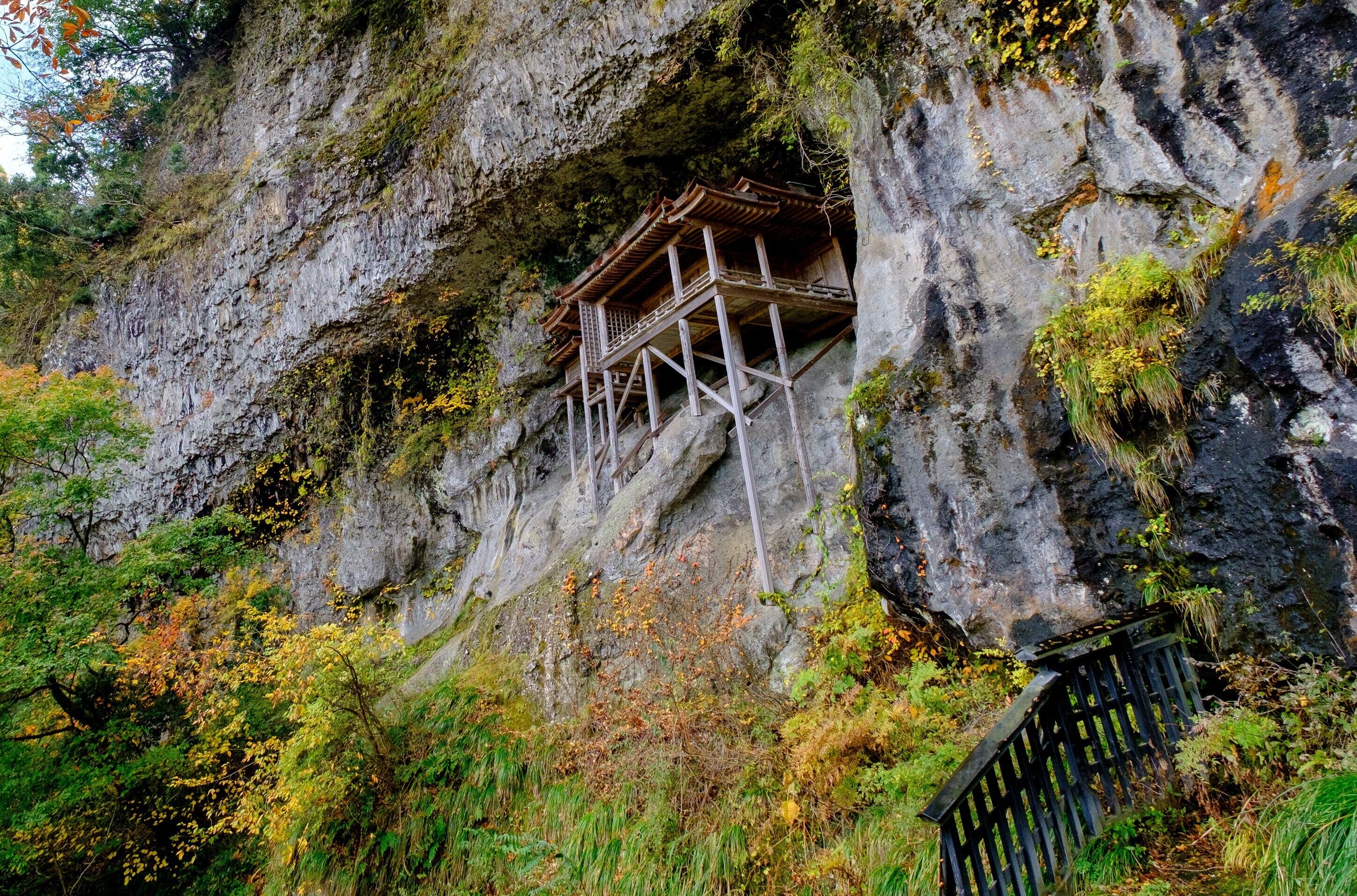 Uma estrutura de madeira tradicional construída na encosta de um penhasco, rodeada por vegetação exuberante e folhagem outonal.