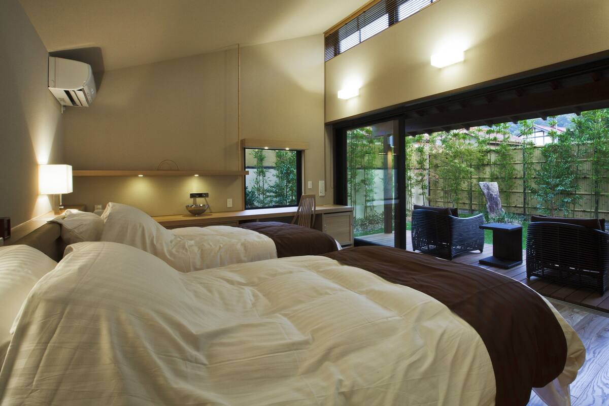 A hotel room with two beds, a large window, and a view of greenery outside.