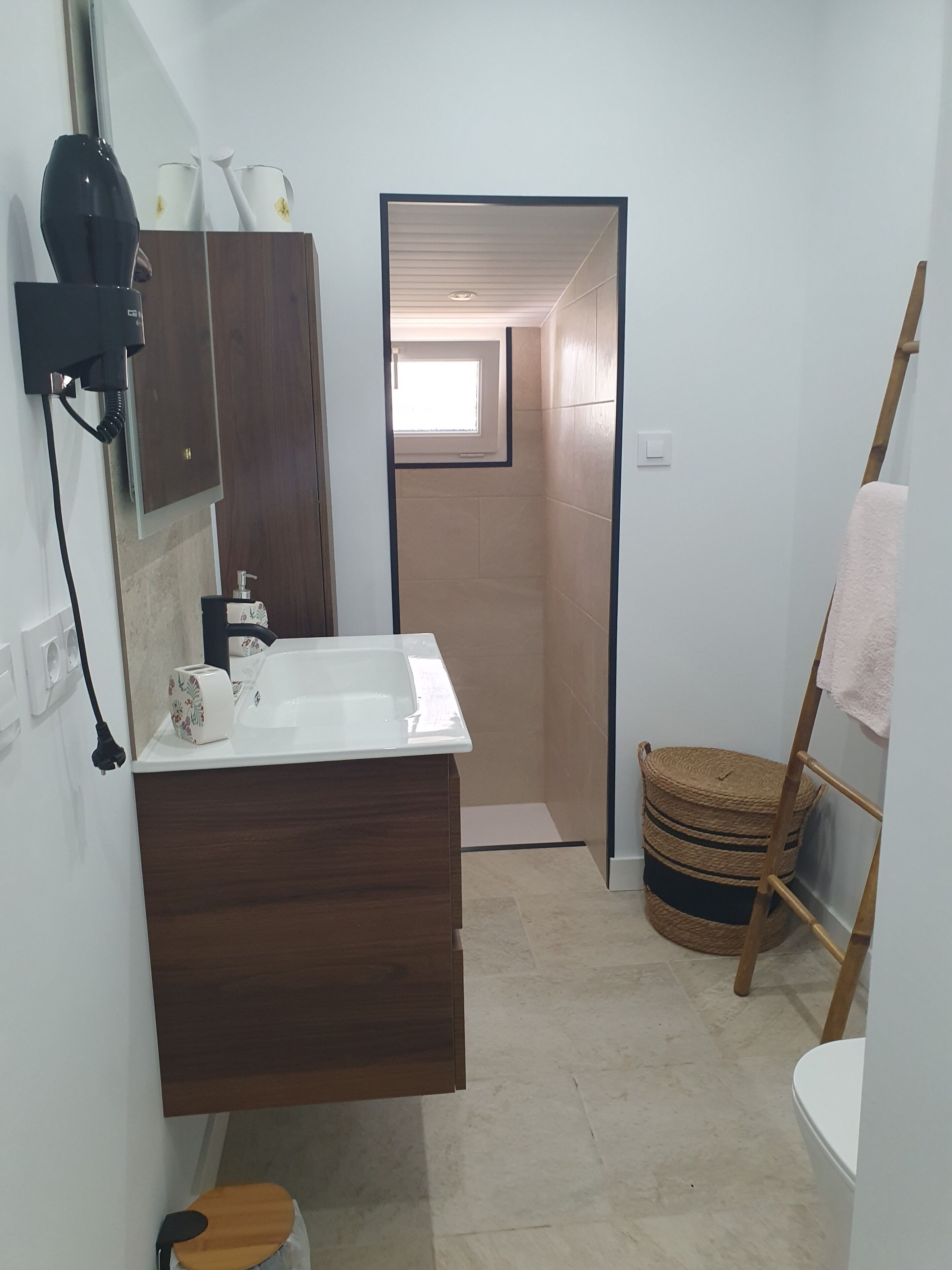Une salle de bain avec un meuble-lavabo en bois, un lavabo blanc, un miroir, un sèche-cheveux, un porte-serviettes et un petit tabouret.