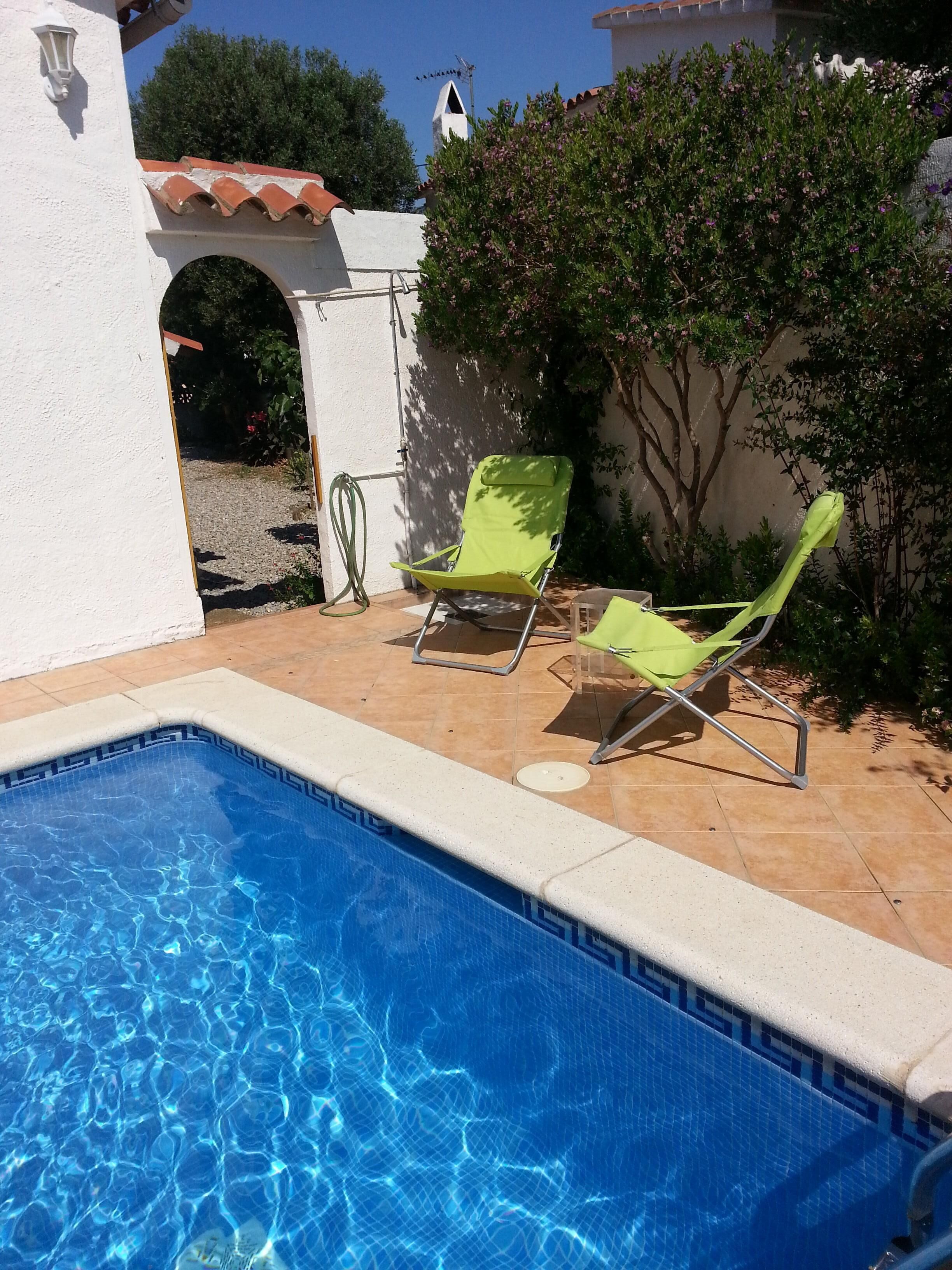 Une piscine bordée de deux chaises longues vertes et d’une petite table, le tout entouré d’un mur blanc et de verdure.