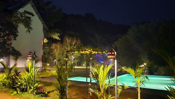 A nighttime view of a resort with a swimming pool, a white building with a sloped roof, and string lights. - Vythiri Greens Resort (Vythiri)