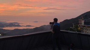 A person with a camera on a balcony overlooking a coastal landscape with hills and a sunset. - Box Inn Jiufen - Hostel (New Taipei City)
