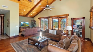 A spacious living room with a high ceiling, wooden beams, a large window, and a view of the outdoors.