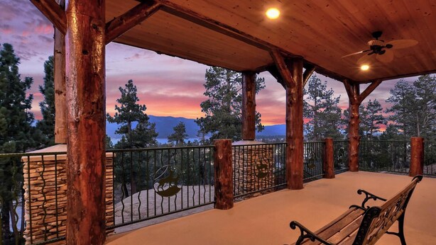 Una terraza de madera con un banco, pared de piedra y barandilla metálica con vistas a un paisaje montañoso.