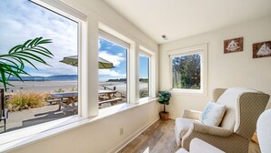 Een kamer met grote ramen met uitzicht op een strand, een houten vloer en een zithoek met een bank en stoelen.