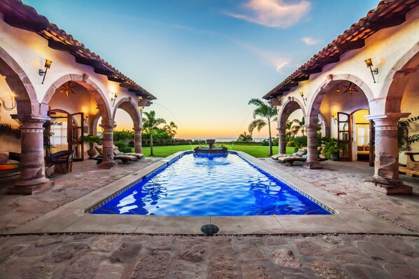 A pool with a fountain, surrounded by arches and columns, with a view of the sunset.
