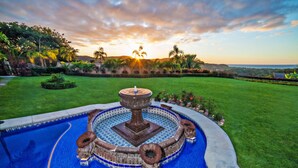 Una piscina con una fuente, rodeada de césped cuidado y plantas tropicales, con un atardecer de fondo.