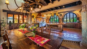 Un comedor rústico con mesa y sillas de madera, una araña y vistas a la cocina y al jardín a través de ventanales arqueados.