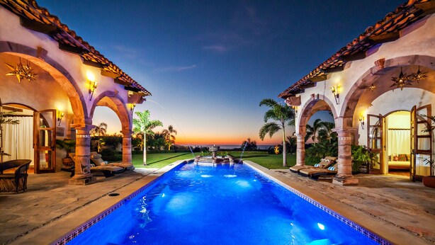 Una piscina con borde de azulejos, rodeada de arcos y columnas, con un atardecer de fondo.