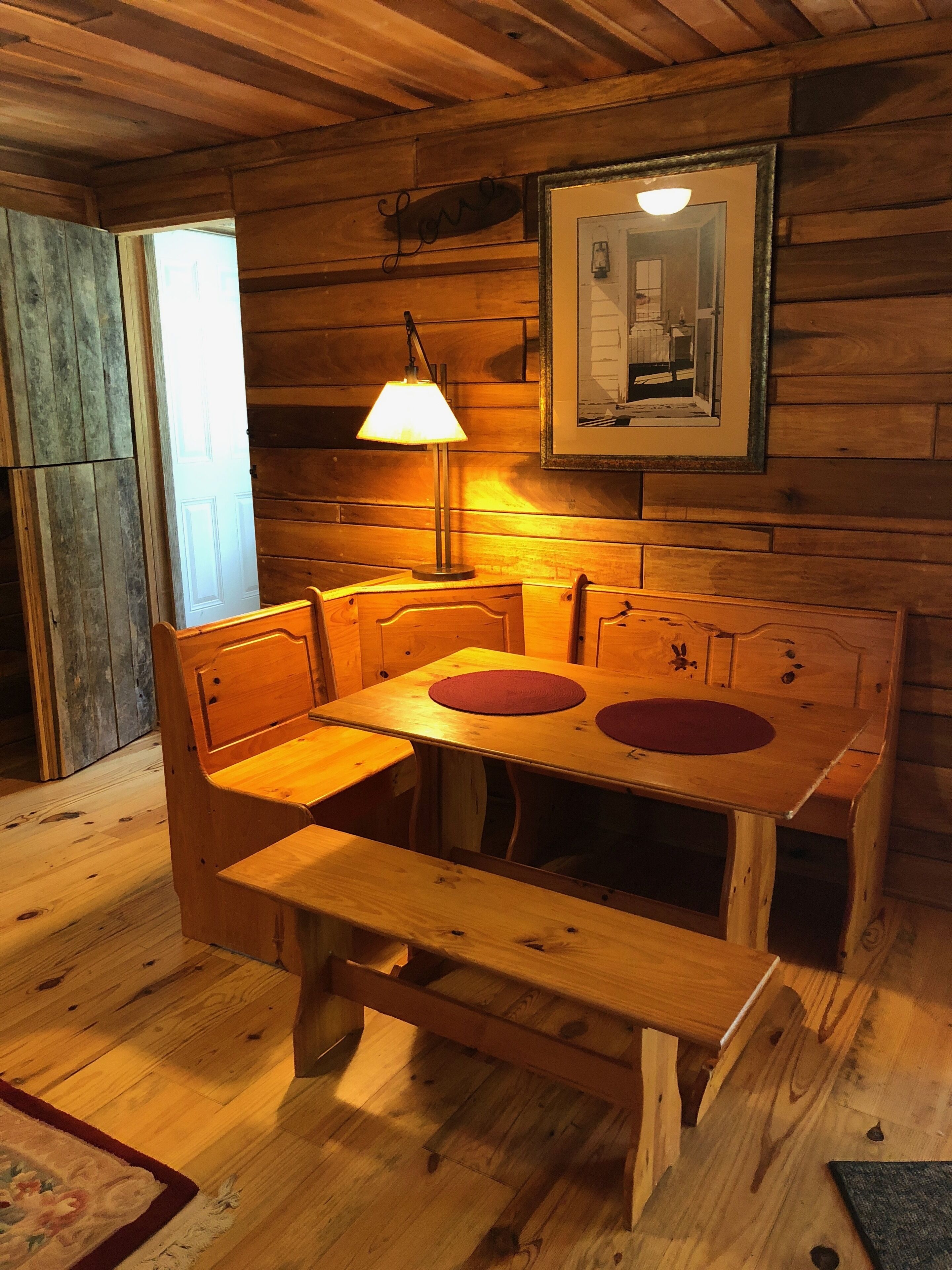 Une table à manger en bois, avec des bancs, une lampe et un tableau encadré accroché au mur.