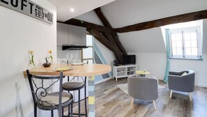 Een loft-achtige kamer met een houten eettafel, twee stoelen, een televisie op een standaard en een groot raam.