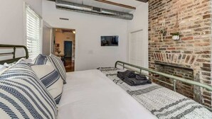 Un dormitorio con pared de ladrillo, una cama con ropa de cama a rayas, una mesita pequeña y una planta colgante.
