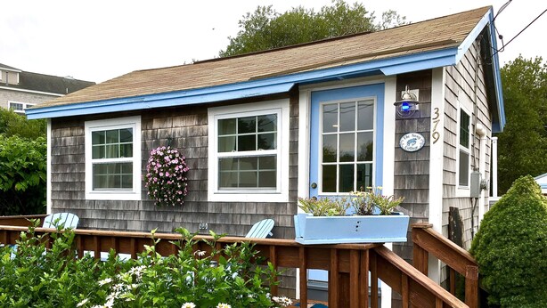 A wooden house with a blue door and windows, a porch with chairs, and a flower bed in front.