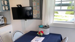 A dining area with a table set for two, a television mounted on the wall, and a window with a view of greenery outside.