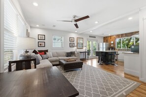 A modern living room with a sofa, ottoman, dining table, and kitchen area with a refrigerator and cabinets.
