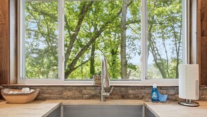Une cuisine avec une grande fenêtre, un évier en acier inoxydable, un saladier en bois et une bouteille de liquide vaisselle.